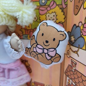 May include: A doll wearing a white top and pink skirt holding a small, white pillow with a brown teddy bear illustration and a pink bow. The background features a colorful, cartoon-style design with various elements.