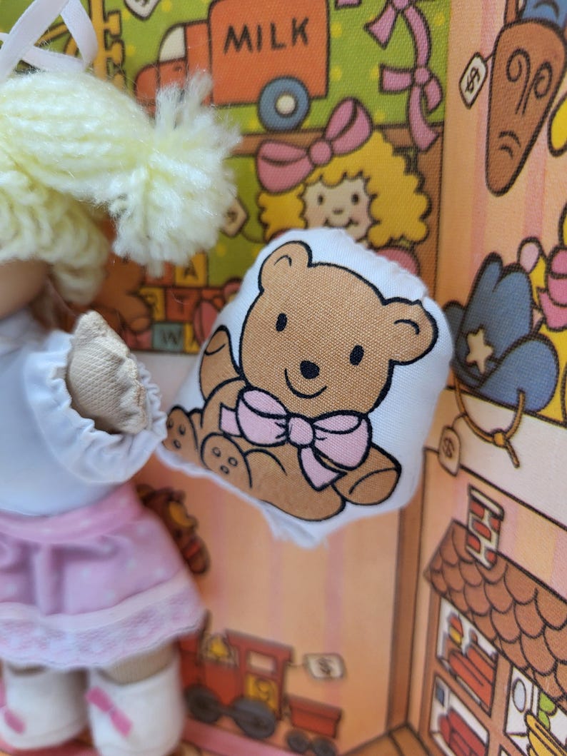 May include: A doll wearing a white top and pink skirt holding a small, white pillow with a brown teddy bear illustration and a pink bow. The background features a colorful, cartoon-style design with various elements.