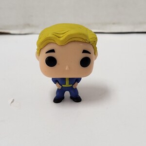 Puede incluir: Figura de vinilo de un personaje con peinado rubio, ojos negros y un mono azul con un emblema amarillo. La figura está de pie con las manos juntas al frente. La figura es un objeto de colección.
