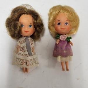 May include: Two vintage doll figures. One doll has brown hair and wears a lace dress with a gray ribbon. The other doll has blonde hair and a purple dress with a flower detail. Both dolls have blue eyes and rosy cheeks.