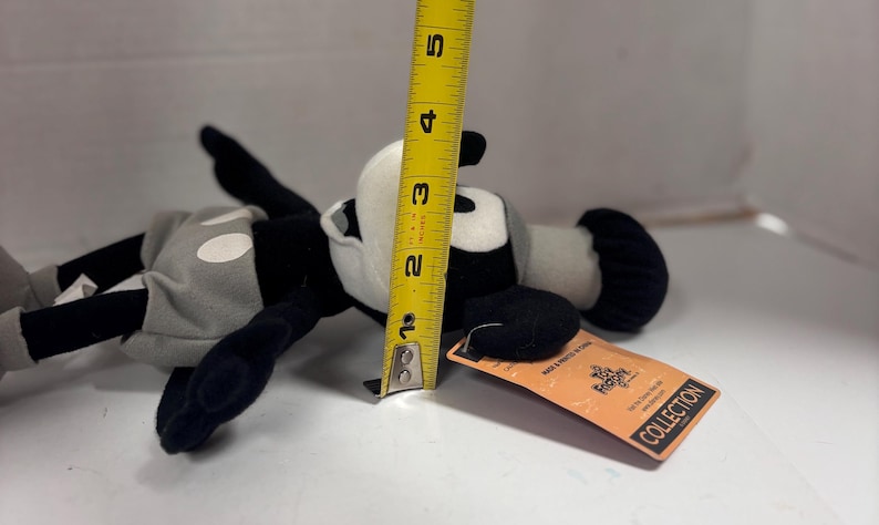 May include: A black, white, and gray plush toy, approximately 4 inches tall, of a cartoon character. The toy has a tag attached. The character is lying on its side.