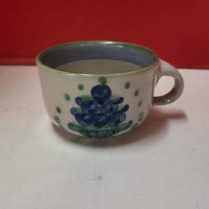 Puede incluir: Una taza de cerámica blanca con un diseño floral azul y verde. La taza tiene un asa y una forma redondeada.