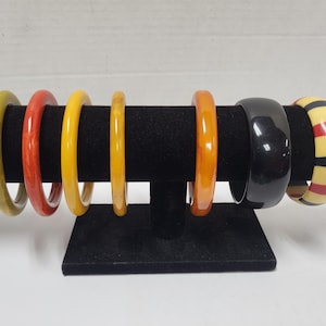 May include: A collection of seven colorful bangles displayed on a black stand. The bangles vary in color, including green, red, yellow, orange, black, and a striped design of red, yellow, and black. The bangles are made of a glossy material.