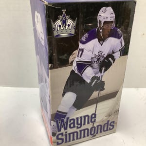 May include: A rectangular cardboard box featuring a graphic of hockey player Wayne Simmonds in a white, purple, and black Los Angeles Kings uniform. The box displays the team logo, the player's name in bold text, and a depiction of him holding a hockey stick.