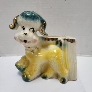 May include: A vintage ceramic dog planter. The dog is white with yellow and green accents, a blue head, and gold trim. The planter is shaped like a tree stump.