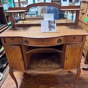 May include: A wooden sideboard featuring a central open shelf, two side cabinets with doors, and a top mirror with two small shelves. The piece has curved legs, metal hardware, and a rectangular information card placed on the surface.