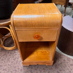 May include: A light brown wooden nightstand featuring a curved top edge, a single drawer with a round orange knob, and an open storage compartment at the bottom. The piece has a smooth finish and a rectangular base with horizontal detailing.