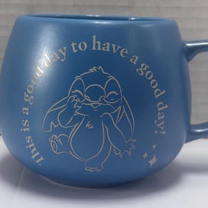 May include: Blue ceramic mug with a rounded shape and a handle. The mug features a cartoon character illustration and the text "This is a good day to have a good day!" in a lighter blue color.