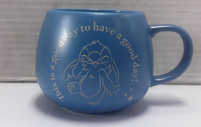 May include: Blue ceramic mug with a rounded shape and a handle. The mug features a cartoon character illustration and the text "This is a good day to have a good day!" in a lighter blue color.