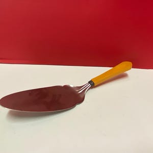 May include: A silver cake server with a yellow plastic handle. The server has a slightly curved blade and a rounded tip.