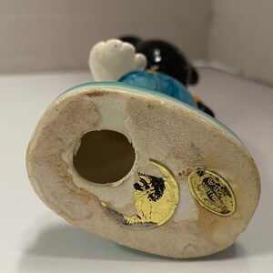 May include: Ceramic figurine of Mickey Mouse wearing blue overalls and a yellow shirt. The figurine is standing on a blue base with a gold sticker that reads "Disney Traditions by Enesco".