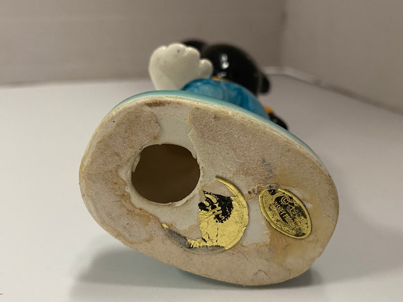May include: Ceramic figurine of Mickey Mouse wearing blue overalls and a yellow shirt. The figurine is standing on a blue base with a gold sticker that reads "Disney Traditions by Enesco".