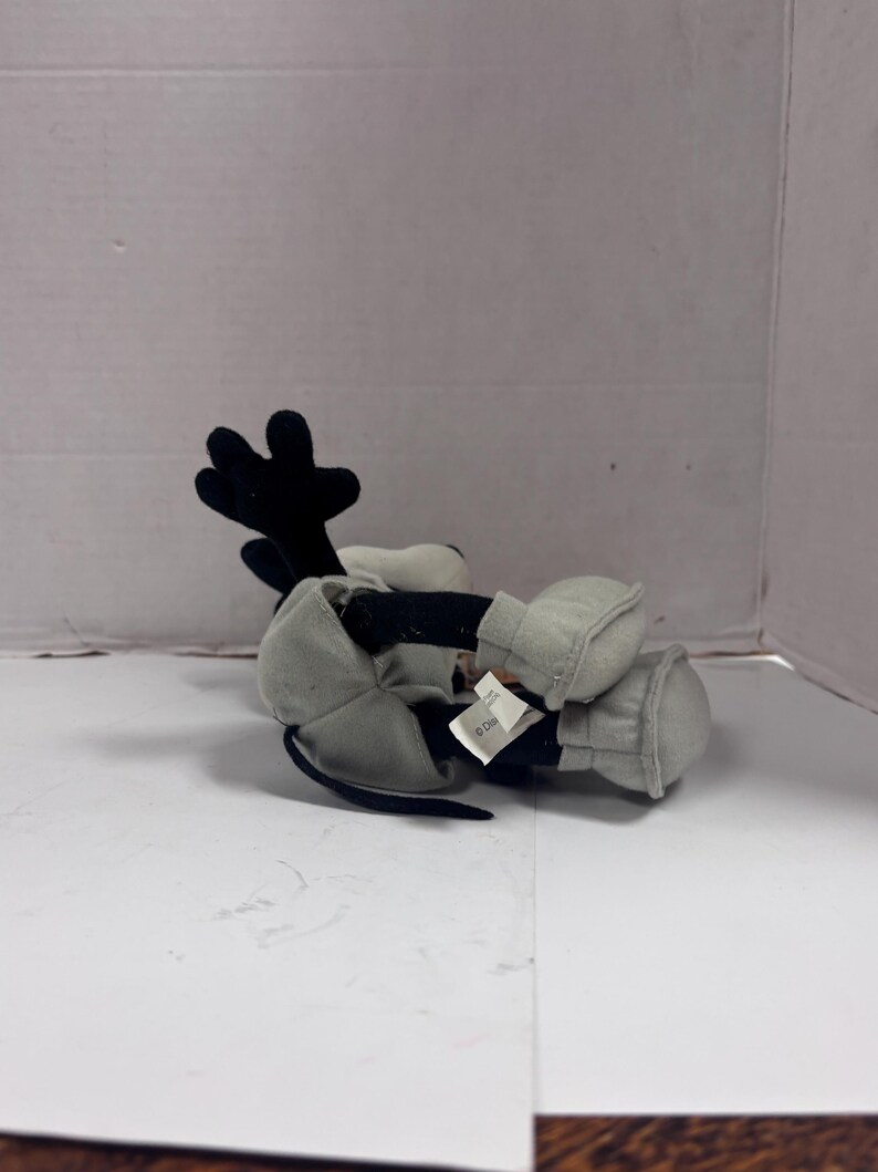 May include: A plush toy of a cartoon character, primarily black and white with gray accents. The character has large black ears, a black tail, and gray gloves and shoes. The toy is lying on its side.