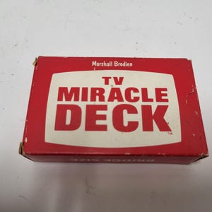 以下が含まれることがあります： 大きな白い文字で「TV MIRACLE DECK」と書かれた赤い箱。箱の上部には「Marshall Brodien」の文字もあります。この箱はブリッジサイズのトランプ用です。
