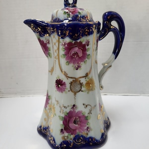Op de afbeelding: Een witte porseleinen koffiepot met een deksel, met een donkerblauwe rand en handvat. De pot is versierd met bloemmotieven in roze, paarse en gele tinten, geaccentueerd met gouden randen. De pot is ongeveer 20 cm hoog.