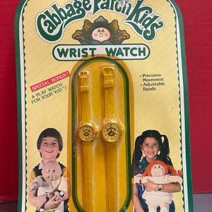 May include: A package for a Cabbage Patch Kids wrist watch. The package features two yellow watches with a Cabbage Patch Kid face on the watch face. The package also features two children holding Cabbage Patch Kids dolls.