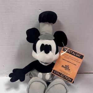 May include: A plush Mickey Mouse toy wearing a chef's hat and outfit. The toy is primarily black, white, and gray, with a tag that reads "NOSTALGIC COLLECTION" and "MADE & PRINTED IN CHINA".