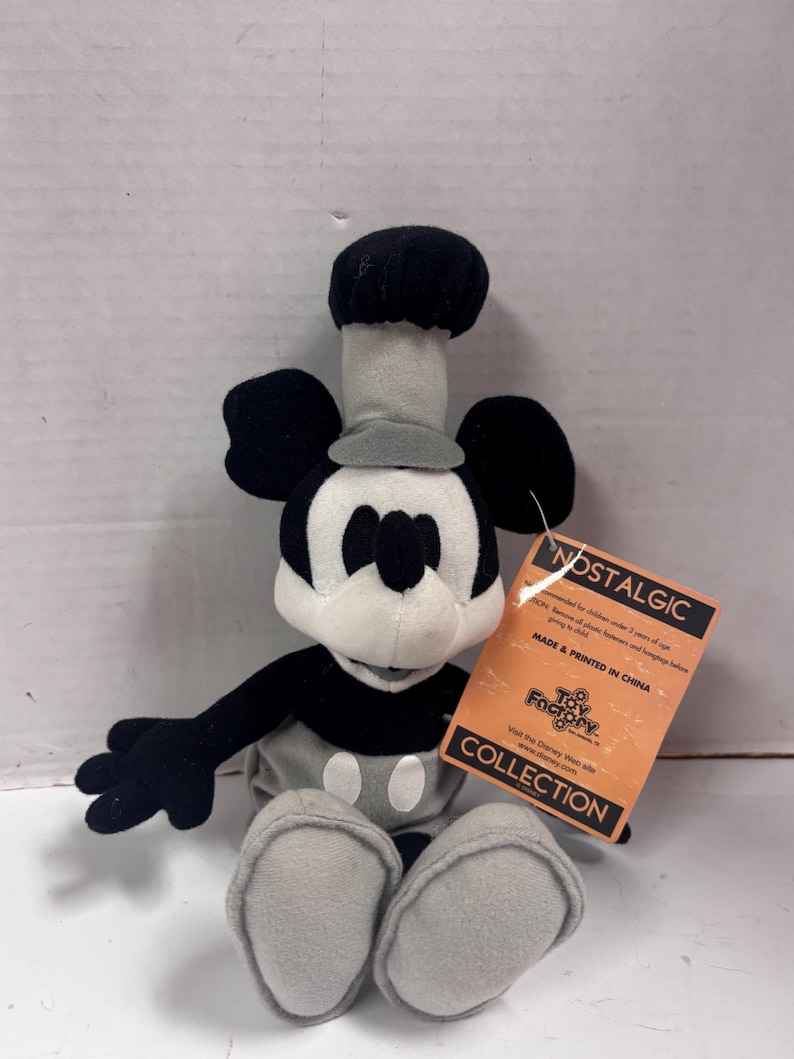 May include: A plush Mickey Mouse toy wearing a chef's hat and outfit. The toy is primarily black, white, and gray, with a tag that reads "NOSTALGIC COLLECTION" and "MADE & PRINTED IN CHINA".