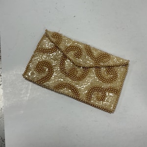 May include: A rectangular, cream-colored beaded clutch with a flap closure. The clutch features a swirling pattern of gold-colored beads, creating a textured surface. The edges are also lined with gold beads.