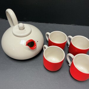 Puede incluir: Un juego de té de cerámica que consta de una tetera redonda de color blanco roto con el pico y la tapa bordeados en rojo, acompañado de cuatro tazas a juego. Cada taza presenta un exterior rojo brillante y un interior blanco roto.