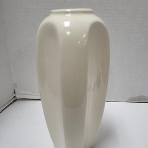 Puede incluir: Un jarrón de cerámica color crema con un diseño acanalado. El jarrón tiene un cuerpo ancho y redondeado que se estrecha hacia la base y una abertura ligeramente ensanchada. La superficie es lisa y brillante, reflejando la luz.