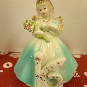May include: A porcelain figurine of a girl in a blue dress holding a bouquet of flowers. The girl is wearing a crown and has the number 16 on her dress.