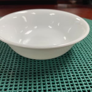 以下が含まれることがあります： 滑らかで光沢のある白いセラミックボウル。ボウルは空で、緑色の模様の表面の上に置かれています。