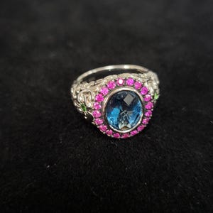 May include: A silver-toned ring featuring an oval-cut blue center stone surrounded by a halo of small, round, vibrant pink stones. The metal band displays intricate, raised scrollwork patterns along the sides, accented with tiny green stones.