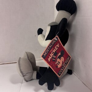 May include: A plush Mickey Mouse toy in a seated position, featuring black, white, and gray colors. The toy has a black hat, ears, and gloves. A tag reads "NOSTALGIC MICKEY COLLECTION" with a Mickey Mouse graphic.