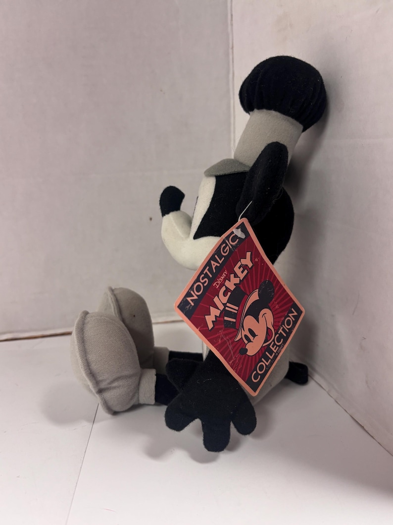 May include: A plush Mickey Mouse toy in a seated position, featuring black, white, and gray colors. The toy has a black hat, ears, and gloves. A tag reads "NOSTALGIC MICKEY COLLECTION" with a Mickey Mouse graphic.