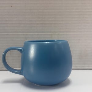 May include: A solid blue mug with a rounded body and a curved handle. The mug is a light shade of blue and sits on a white surface. The background is a light gray.