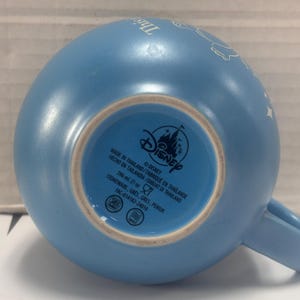 May include: A light blue Disney ceramic mug. The bottom of the mug features the Disney logo and text indicating it is stoneware, made in Thailand, and holds 21 oz. The mug has a handle and a curved shape.