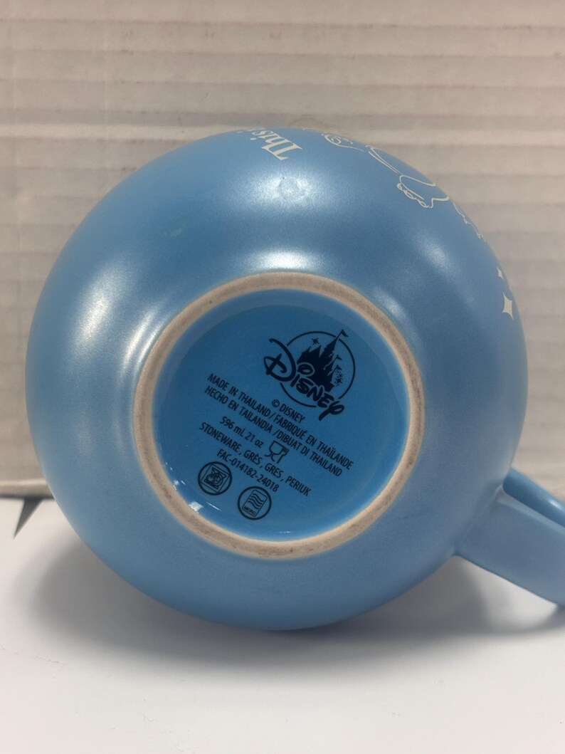 May include: A light blue Disney ceramic mug. The bottom of the mug features the Disney logo and text indicating it is stoneware, made in Thailand, and holds 21 oz. The mug has a handle and a curved shape.