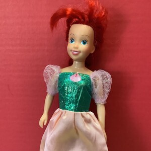 May include: A red-haired doll wearing a green and pink dress with a seashell design. The doll has a smiling face with blue eyes.