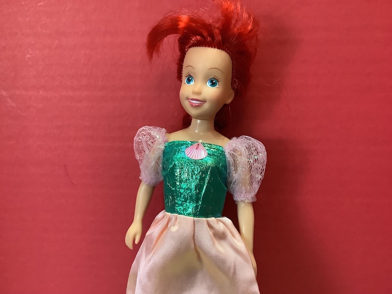 May include: A red-haired doll wearing a green and pink dress with a seashell design. The doll has a smiling face with blue eyes.