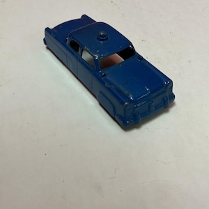 May include: A vintage die-cast metal toy car in a deep blue color. The classic car has a detailed design with visible wear and tear, suggesting it's an older model. The car features a roof light and open windows.