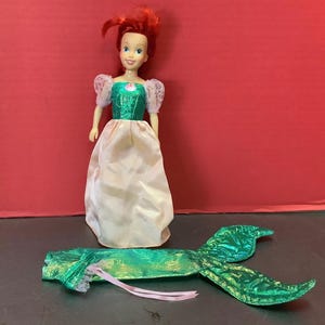 May include: A red-haired doll wearing a green and white dress with a pink sash. The doll is holding a green and gold mermaid tail.