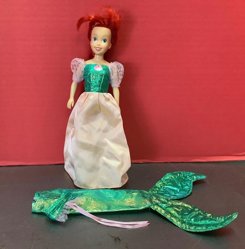 May include: A red-haired doll wearing a green and white dress with a pink sash. The doll is holding a green and gold mermaid tail.