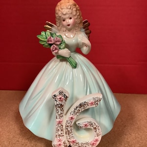 May include: A porcelain figurine of a girl in a light blue dress with a gold halo and wings. She is holding a bouquet of pink flowers and the number 16 is on the base of the figurine.
