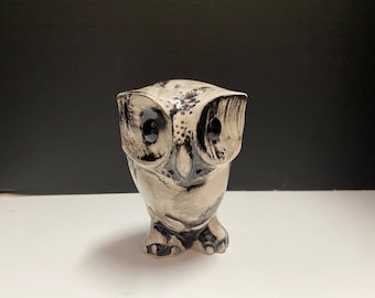 May include: A ceramic owl figurine with black and white painted details. The owl has large black eyes and a detailed beak.