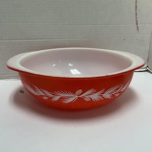 May include: A round, orange glass bowl with a white interior and integrated side handles. The exterior features a repeating white graphic pattern of pine branches and pinecones encircling the middle of the bowl in a horizontal band.