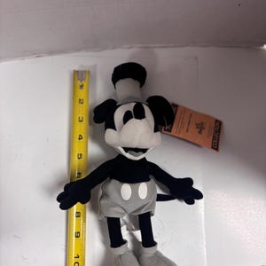 May include: A plush Mickey Mouse toy, primarily black and white with gray shorts and shoes. The toy is approximately 11 inches tall. It has a tag attached. The classic cartoon character is in a standing pose.