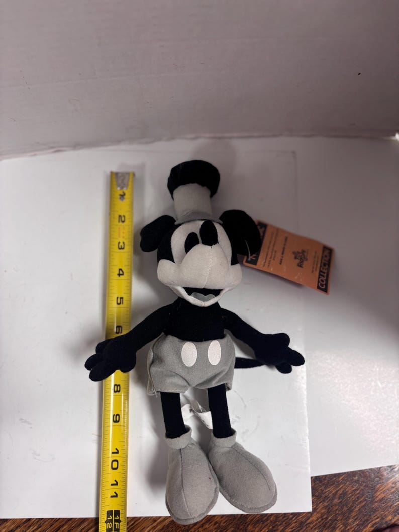 May include: A plush Mickey Mouse toy, primarily black and white with gray shorts and shoes. The toy is approximately 11 inches tall. It has a tag attached. The classic cartoon character is in a standing pose.