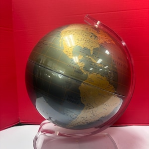 Może przedstawiać: Globus w kolorze ciemnoszarym i złotym, zamontowany na przezroczystym, plastikowym stojaku. Globus zawiera szczegóły geograficzne, w tym kontynenty i nazwy krajów. Stojak ma przezroczystą, zakrzywioną konstrukcję.