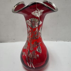 Puede incluir: Un jarrón de cristal rojo rubí con borde plateado ondulado. El jarrón presenta un diseño floral plateado, con flores y tallos detallados. El jarrón tiene una base bulbosa y una parte superior acampanada.