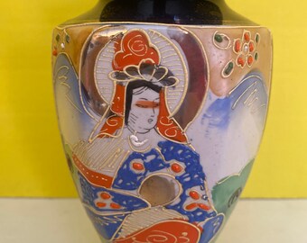 May include: A ceramic vase featuring a dark blue neck and base with a central hand-painted illustration of a figure in traditional attire. The design includes gold-outlined details in orange, blue, and green, set against a cream-colored background.