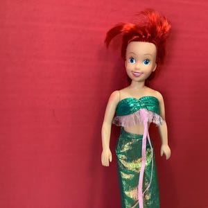 May include: A red-haired doll wearing a green and pink mermaid costume. The doll has blue eyes and a friendly smile.