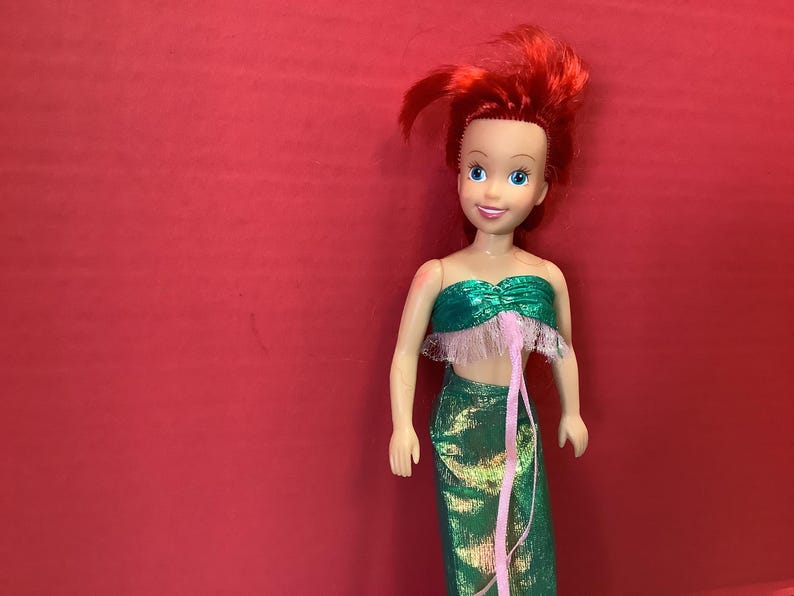 May include: A red-haired doll wearing a green and pink mermaid costume. The doll has blue eyes and a friendly smile.