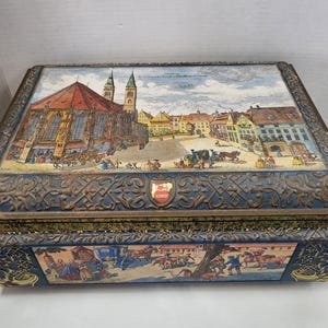 May include: A vintage tin box with a blue and gold design. The lid features a colourful illustration of a European town square with a church in the background. The text "Schmidts" is visible on the box.