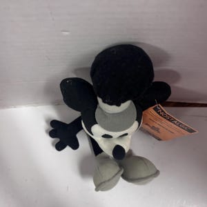 May include: A black and white plush toy mouse with large ears, a round head, and a white face. The mouse has black arms and a tail, with gray shoes. A tag that says "NOSTALGIC" is attached to the toy.